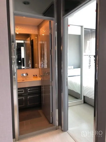 Condominium for rent in Khlong Toei, Bangkok - Hallway showing a modern bathroom with vanity beside the entrance to a bedroom with mirrored wardrobe.