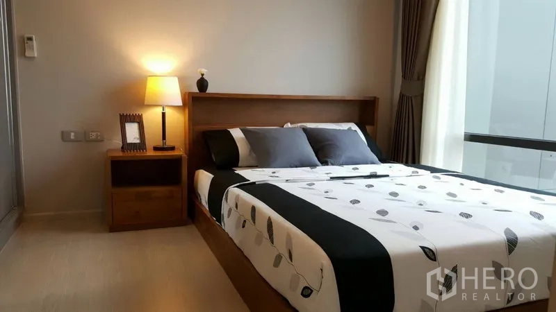 Condominium for rent in Khlong Toei, Bangkok - Cozy bedroom with double bed, bedside lamp and large window with curtains.