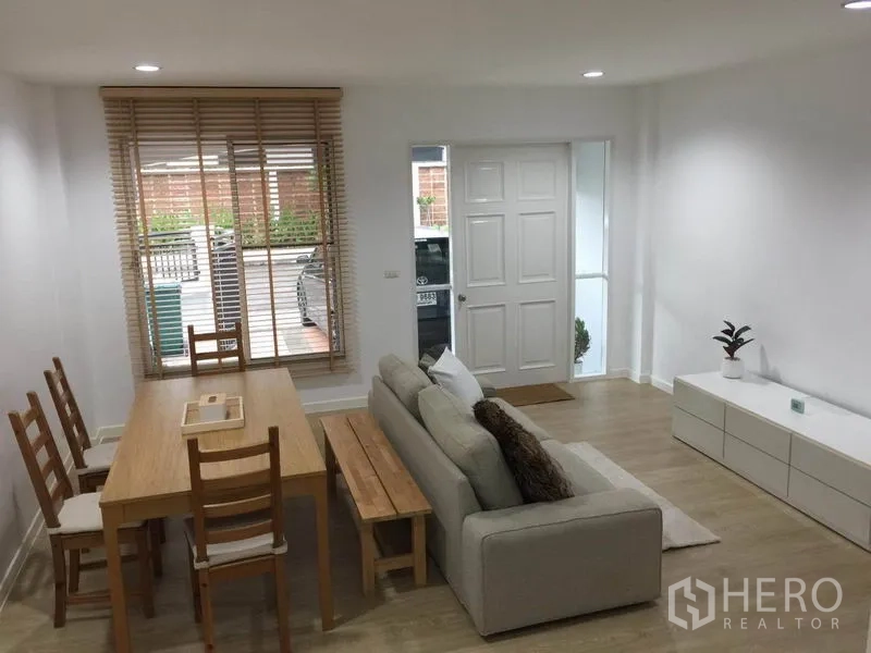 Townhouse for rent in Lat Phrao, Bangkok - Bright open-plan living and dining area with sofa, wooden table and front window.