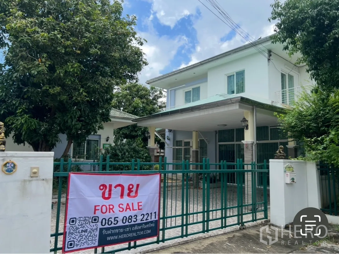 บ้าน ขายในMin Buri, Bangkok - มุมมองหน้าบ้านเดี่ยว 2 ชั้น โครงการบ้านสวนร่มเกล้า พร้อมรั้วสีเขียวและป้ายประกาศขาย