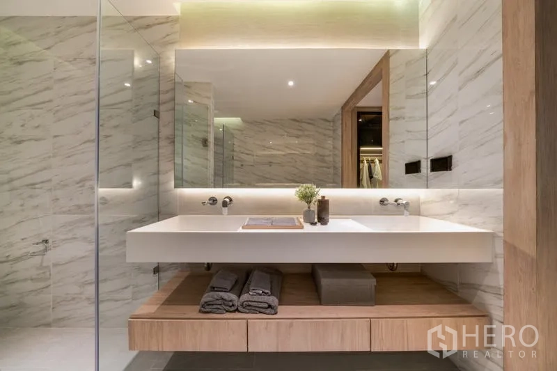 Condominium for rent in Watthana, Bangkok - Close-up of floating vanity with backlit mirror and storage in a marble-finished bathroom.