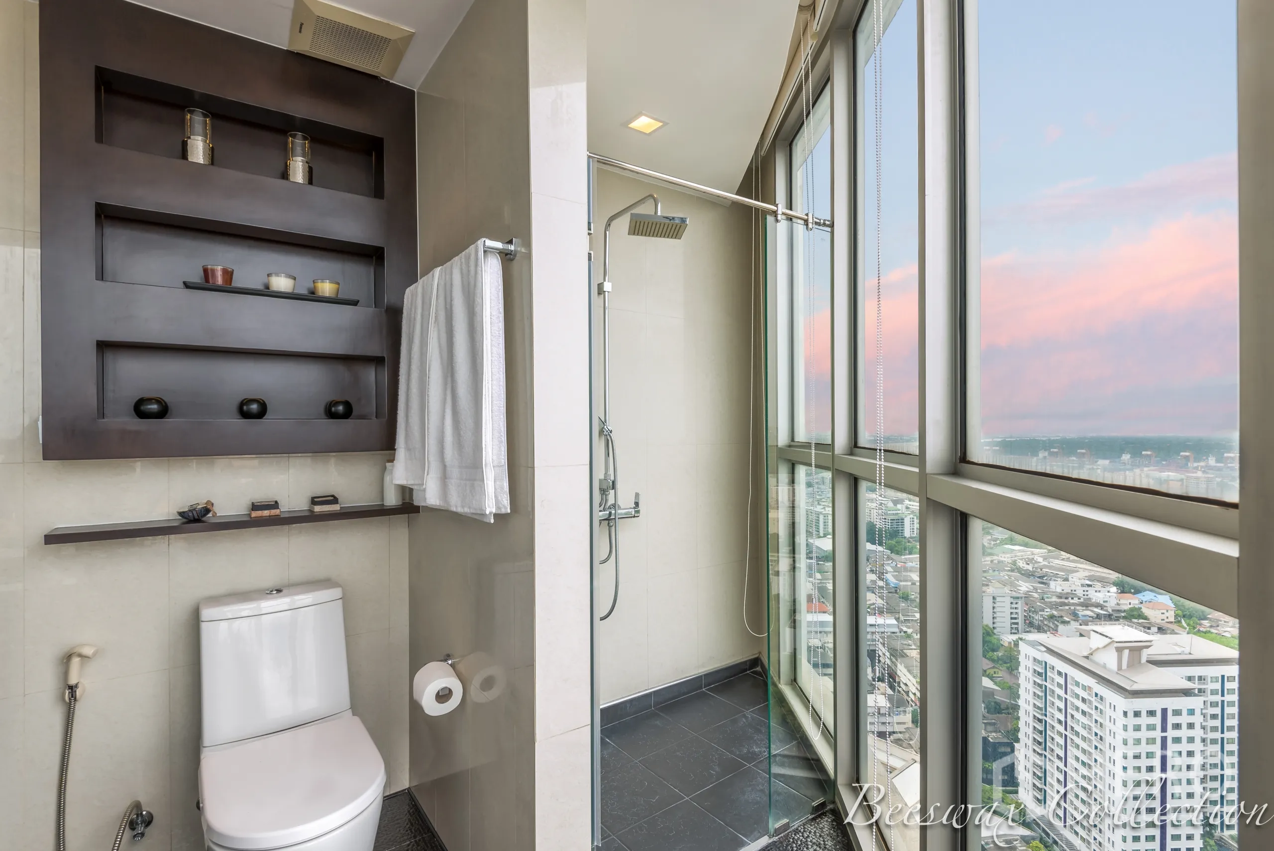 Condominium for rent in Watthana, Bangkok - Bathroom with walk‑in shower beside full‑height windows and built‑in shelving above the toilet, overlooking city views.