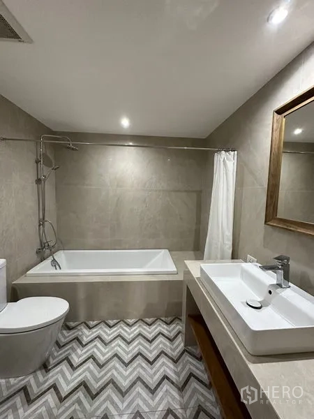 Condominium for rent in Khlong Toei, Bangkok - Second bathroom featuring a bathtub, vanity, toilet and chevron patterned floor tiles.