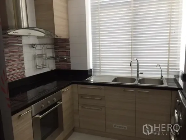 Condominium for rent or sale in Khlong Toei, Bangkok - Modern L-shaped kitchen with oven, range hood, black countertops and a double sink by a large window.
