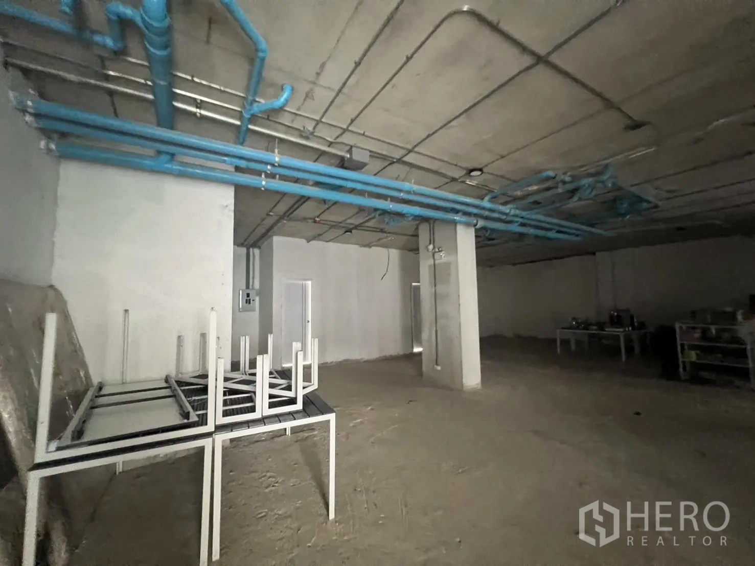 Retail Space for rent - Large open-plan interior with blue ceiling pipes and stacked tables ready for fit-out.