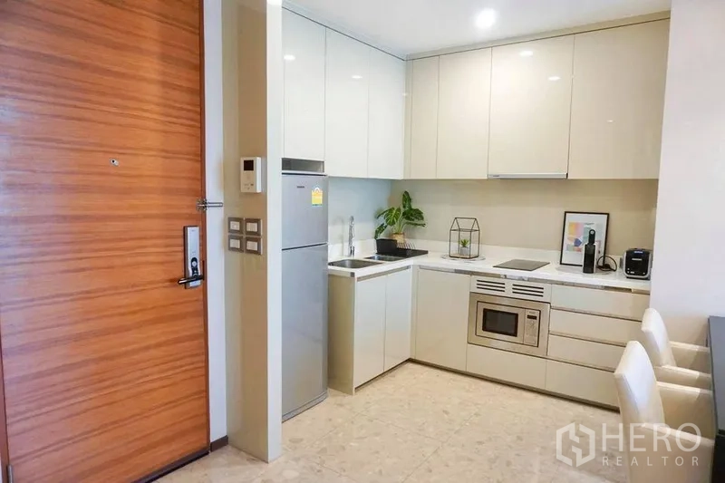 Condominium for rent in Khlong Toei, Bangkok - Compact modern kitchen with glossy cabinets, fridge, cooktop and breakfast seating in Bangkok condo.