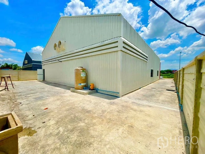 Warehouse for rent in Lam Luk Ka, Pathum Thani - Side view of the warehouse showing a fenced concrete apron and water tank area.