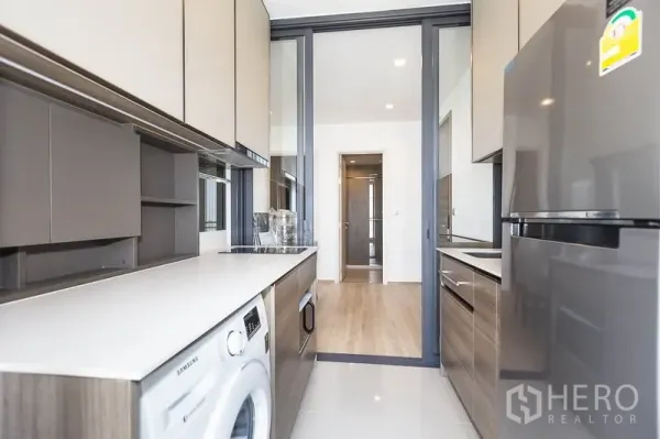 公寓 出租在Phaya Thai, Bangkok - 走廊式厨房，柜体充足，长台面，配冰箱与洗衣机，玻璃推拉门分隔。