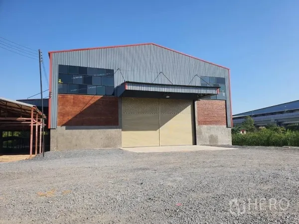 Warehouse for rent in Mueang Pathum Thani, Pathum Thani - Modern steel-clad warehouse exterior with large roller shutter door, gable roof and wide gravel yard in Pathum Thani.