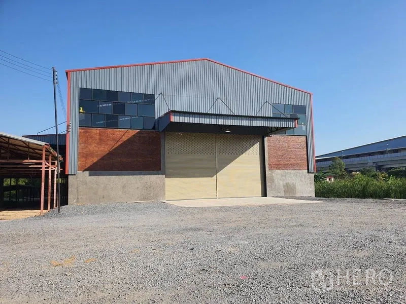 Warehouse for rent in Mueang Pathum Thani, Pathum Thani - Modern steel-clad warehouse exterior with large roller shutter door, gable roof and wide gravel yard in Pathum Thani.