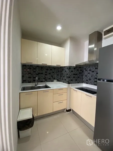公寓 出租在Watthana, Bangkok - L形厨房，米色橱柜、黑色马赛克挡水板，灶台、抽油烟机和冰箱。
