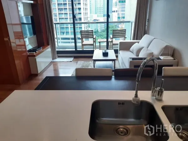 Condominium for rent in Khlong Toei, Bangkok - Open-plan living room with sofa and coffee table leading to a balcony with chairs and Bangkok city view, seen from the kitchen island with double sink.