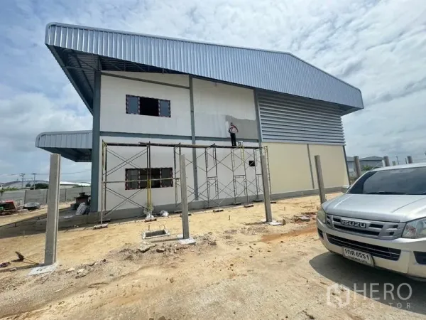 Warehouse for rent in Mueang Samut Prakan, Samut Prakan - Side view of a new steel‑clad warehouse under construction with scaffolding and a wide yard.