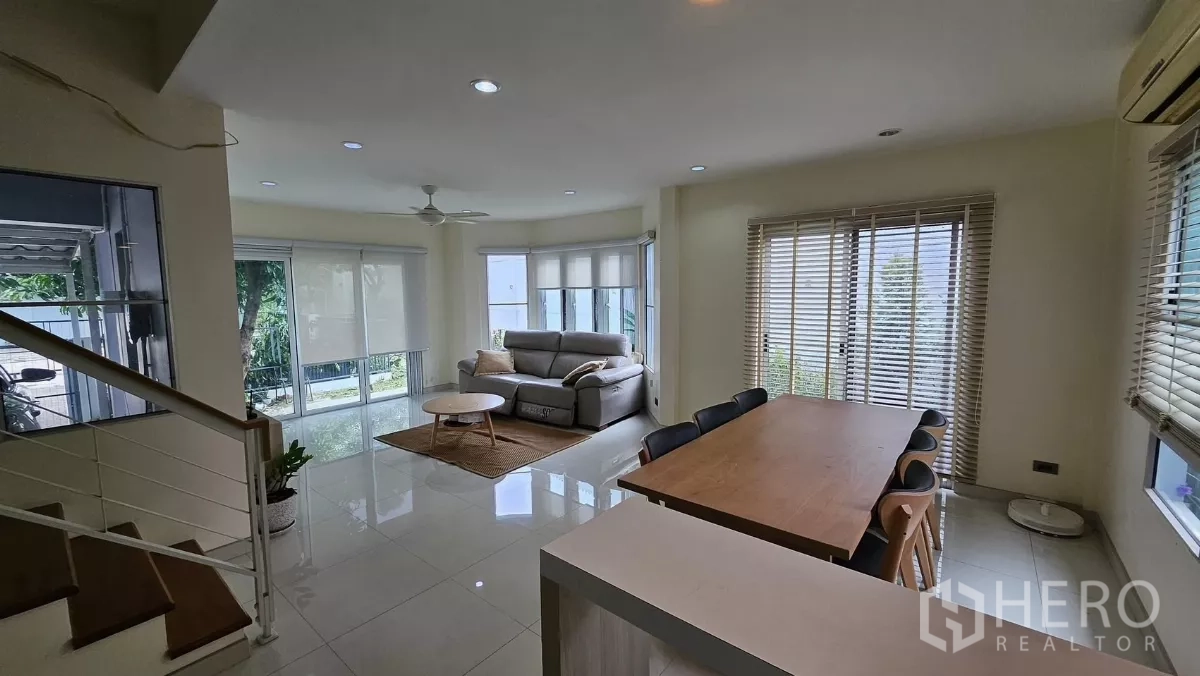 Detached House for rent in Khlong Toei, Bangkok - Spacious living room with corner windows, ceiling fan and adjacent dining table under recessed lights.