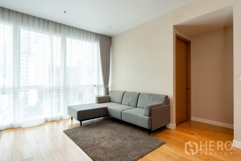 Condominium for rent in Khlong Toei, Bangkok - Bright living room with gray sectional sofa, sheer curtains, and floor-to-ceiling windows over warm wood flooring.