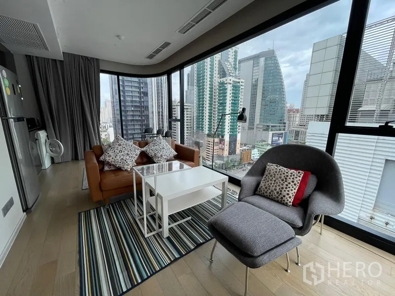 Condominium for rent or sale in Watthana, Bangkok - Corner lounge with wraparound glass walls, city skyline views, sofa, armchair and coffee tables on a striped rug.