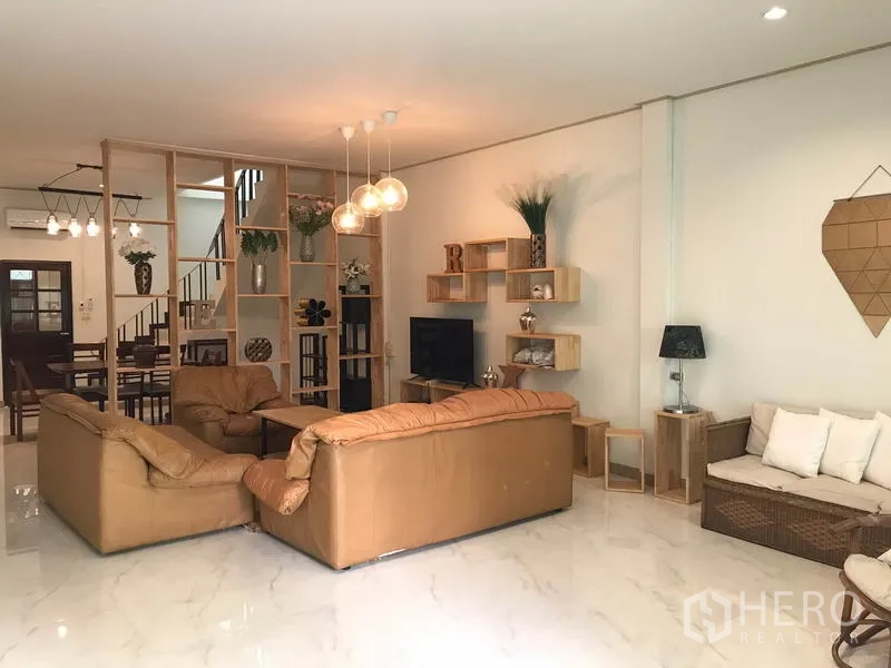 Townhouse for rent or sale in Phra Khanong, Bangkok - Open-plan living room with leather sofas, TV, pendant lights and shelving divider.