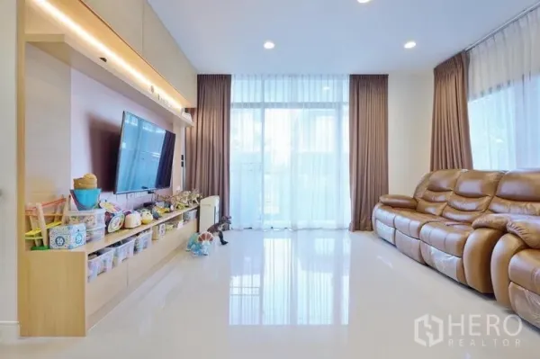 Detached House for rent or sale in Bang Kapi, Bangkok - Sunlit living room with full-height windows, glossy floor, leather sofa and built-in media wall with ample storage.