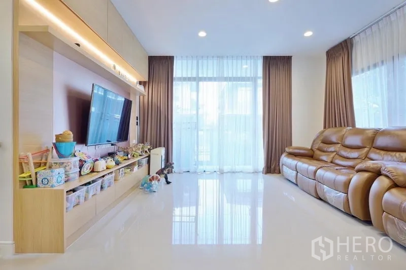 Detached House for rent or sale in Bang Kapi, Bangkok - Sunlit living room with full-height windows, glossy floor, leather sofa and built-in media wall with ample storage.