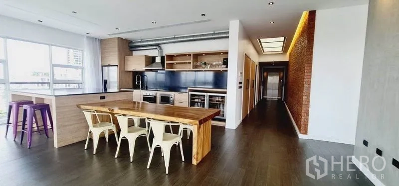 Condominium for rent in Watthana, Bangkok - Open-plan kitchen with wood cabinetry, long dining table, industrial ducting and corridor lighting.