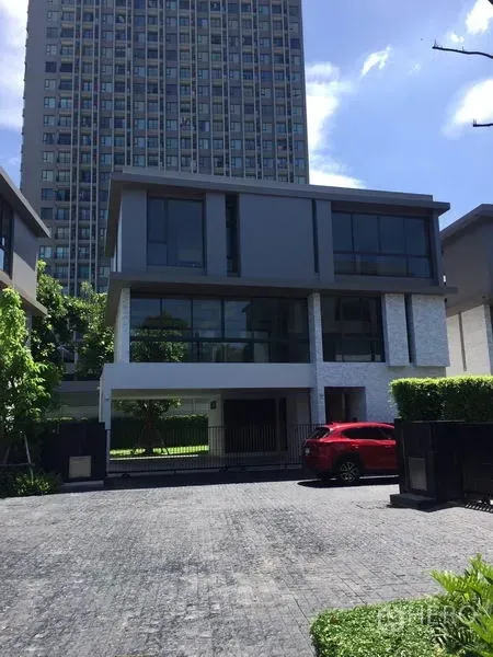 Detached House for rent in Watthana, Bangkok - Contemporary three-storey detached house with floor-to-ceiling windows, gated driveway and a red car, with a high-rise tower behind.