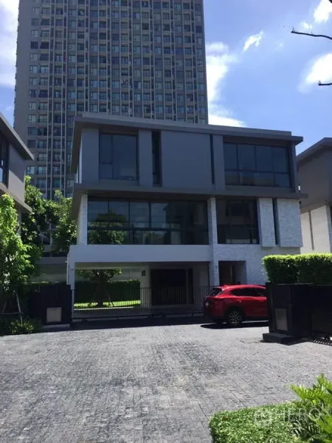 独立式住宅 出租在Watthana, Bangkok - 现代三层独栋，落地窗与封闭车道，前停红色车辆，背景为高层建筑。