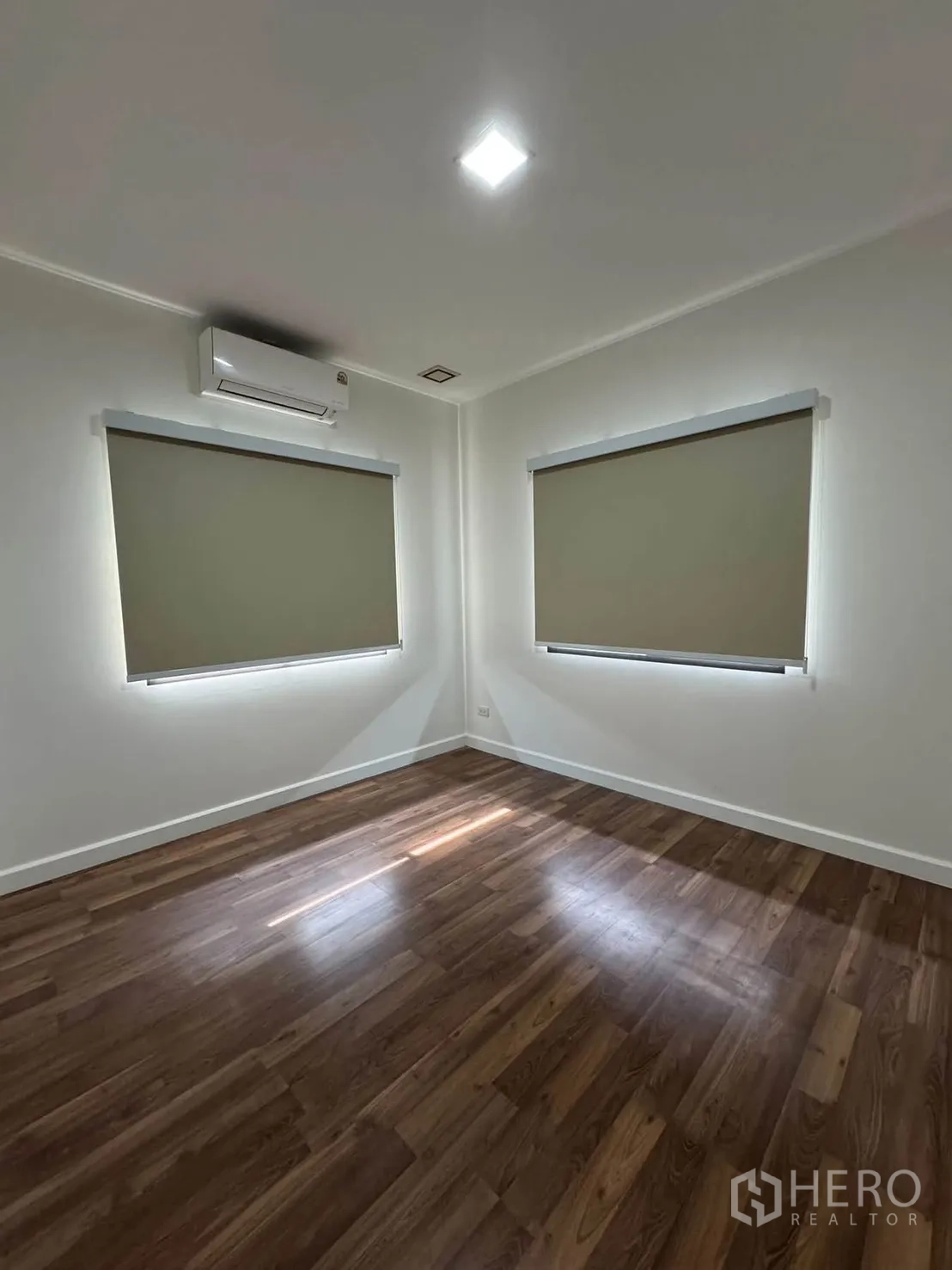 Detached House for rent in Prawet, Bangkok - Empty bedroom with wood flooring, two windows with roller blinds, and an air conditioner.