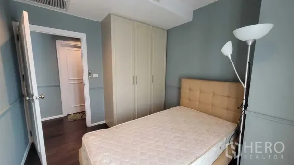 Condominium for rent in Khlong Toei, Bangkok - Bedroom featuring built-in wardrobe, padded headboard and standing lamp against blue walls.