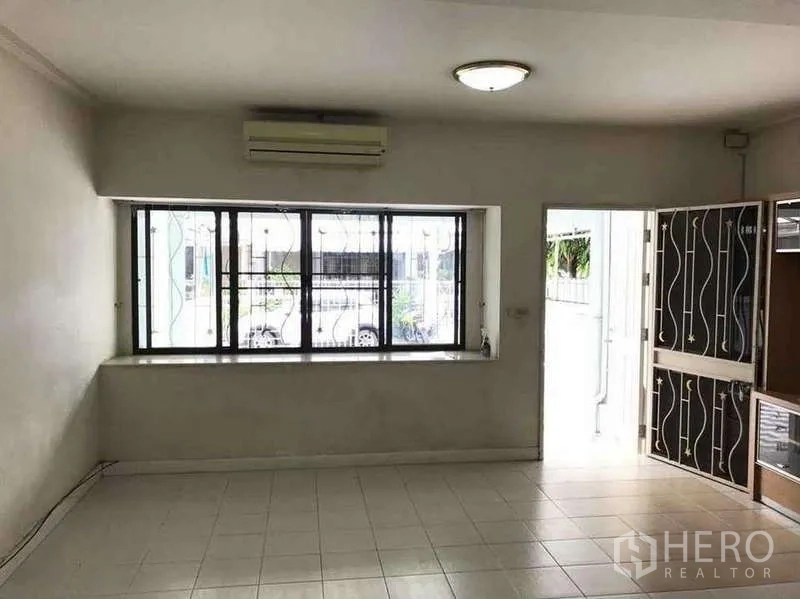 Townhouse for rent in Suan Luang, Bangkok - Front living room with wide window and security bars, air conditioner and door opening to the carport.