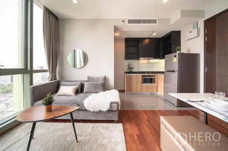 公寓 出租在Ratchathewi, Bangkok - 开放式起居区，灰色沙发与圆镜，背景为厨房与亮面地面。
