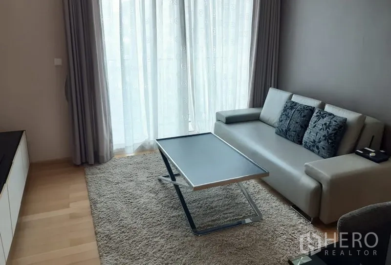 Condominium for rent in Khlong Toei, Bangkok - Bright living room with a grey sofa, slim coffee table, rug, and floor-to-ceiling sheer curtains.