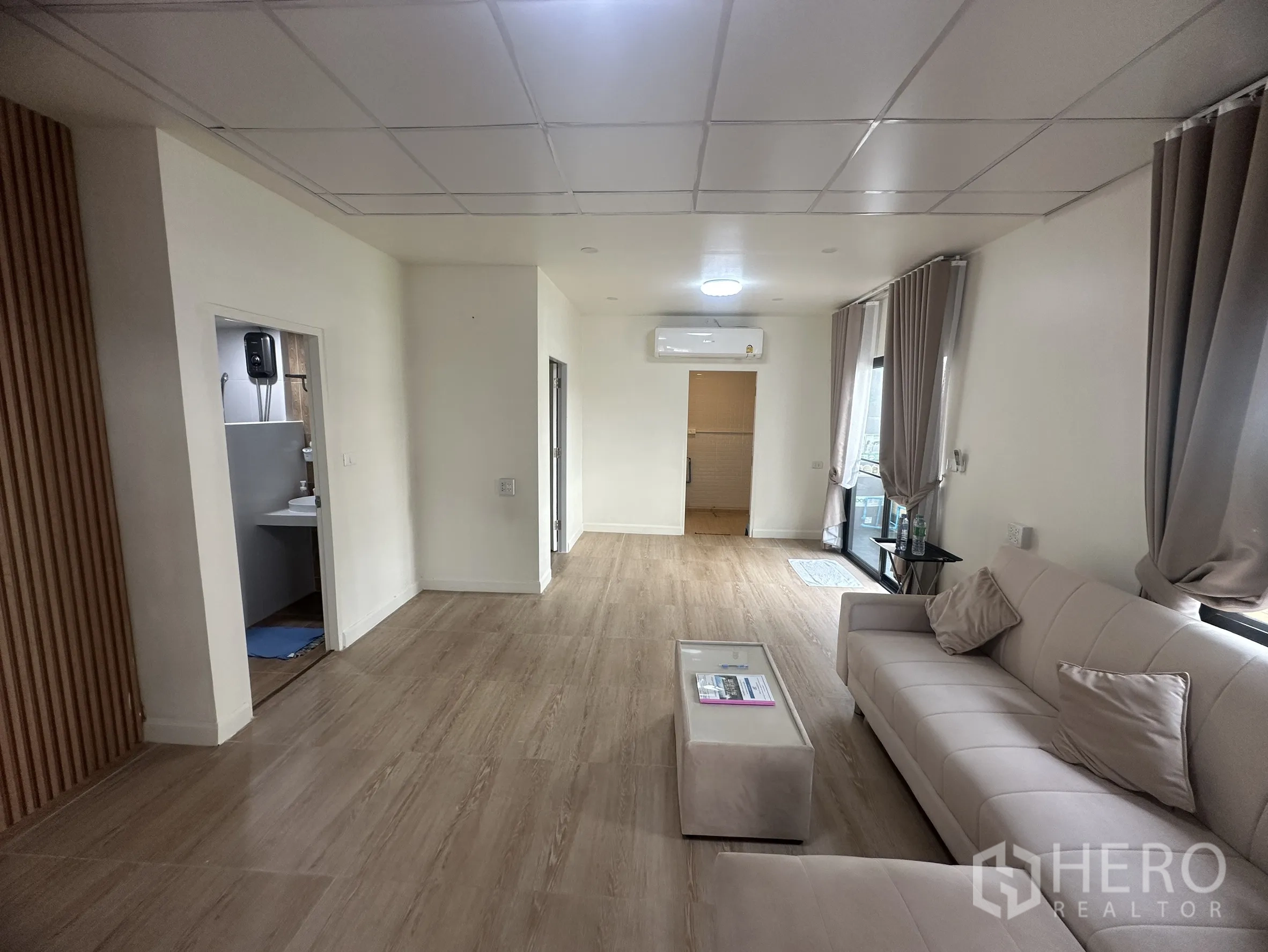 Townhouse for sale in Lam Luk Ka, Pathum Thani - Bright living room with sofa, curtains, wood-look floor tiles, and air conditioner.