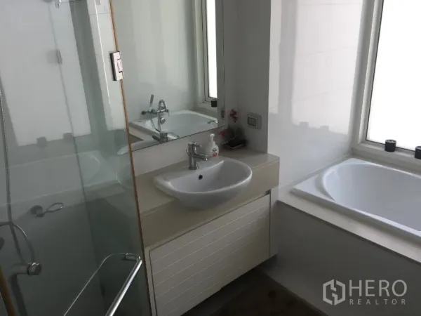 Condominium for rent or sale in Khlong Toei, Bangkok - Contemporary bathroom with glass-enclosed shower, separate bathtub by a window, and a vanity sink.