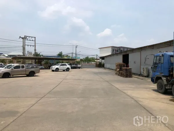 Warehouse for rent in Mueang Pathum Thani, Pathum Thani - Wide concrete yard with cars and trucks beside low-rise warehouse buildings and power lines.