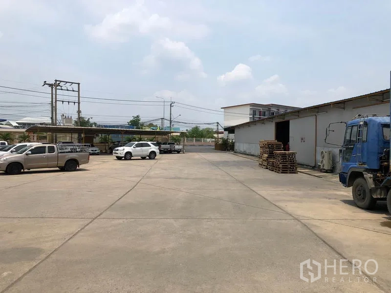 Warehouse for rent in Mueang Pathum Thani, Pathum Thani - Wide concrete yard with cars and trucks beside low-rise warehouse buildings and power lines.