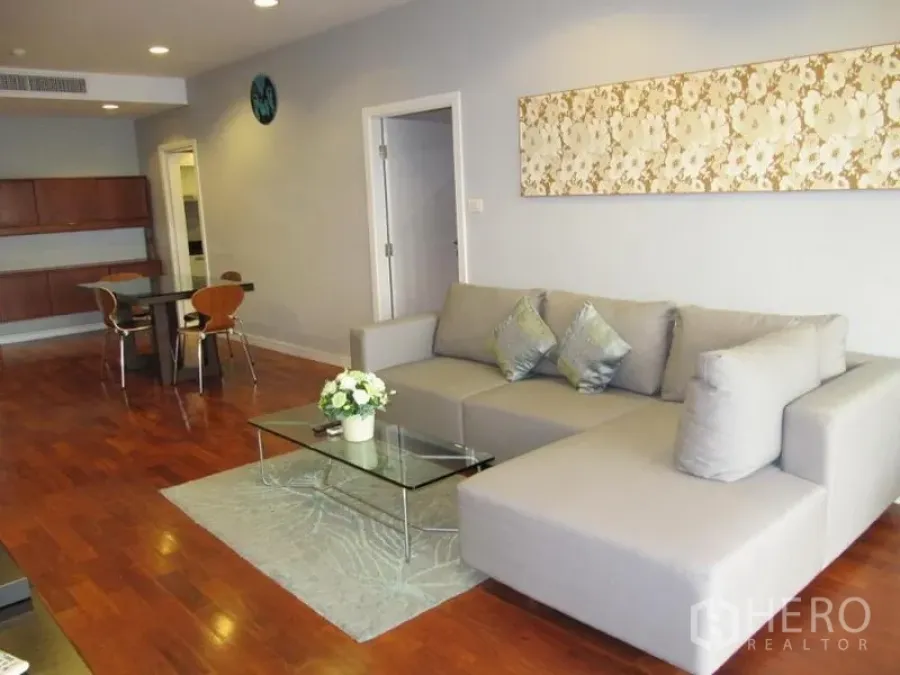 Condominium for rent in Khlong Toei, Bangkok - Open-plan living and dining area with a gray L-shaped sofa, glass coffee table, and warm hardwood floors.