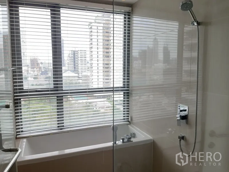 公寓 出租在Watthana, Bangkok - 浴室带浴缸与玻璃淋浴隔断，旁有大窗与百叶，远眺城市天际线。