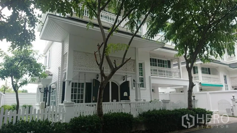 Detached House for rent in Mueang Samut Prakan, Samut Prakan - White three-storey detached house with picket fence, balcony railings and greenery on a quiet lane.