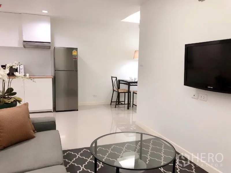 Condominium for rent or sale in Khlong Toei, Bangkok - Open-plan living with wall-mounted TV, round glass coffee table, and compact kitchen.