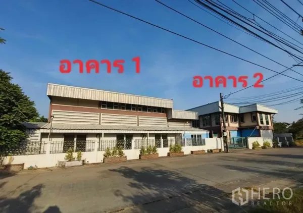 คลังสินค้า ให้เช่าหรือขายในMin Buri, Bangkok - ภาพด้านหน้าถนน เห็นโกดังสองอาคารติดกัน พร้อมรั้วรอบขอบชิดและป้ายระบุอาคาร 1 และอาคาร 2