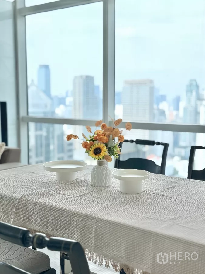 公寓 出租在Pathum Wan, Bangkok - 摆放花艺的餐桌，背后是大幅城市景观窗。