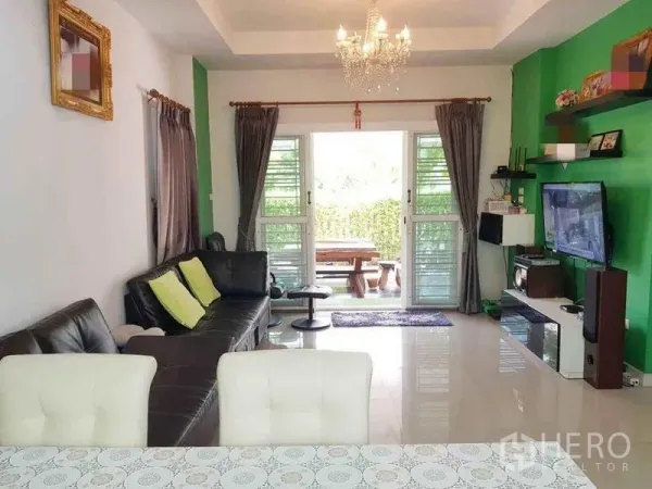 Detached House for sale in Nonthaburi - Bright living room with leather sofa, chandelier, TV on a green accent wall, and sliding doors to a garden patio.