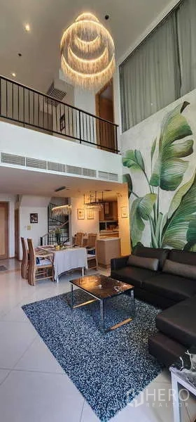 Condominium for sale in Sathon, Bangkok - Double-height living room with mezzanine, L-shaped sofa, tropical wall mural and modern chandelier.