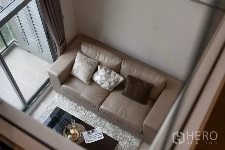 Condominium for rent or sale in Khlong Toei, Bangkok - Top view of a cozy living room with beige leather sofa, rug and sliding doors to the balcony.