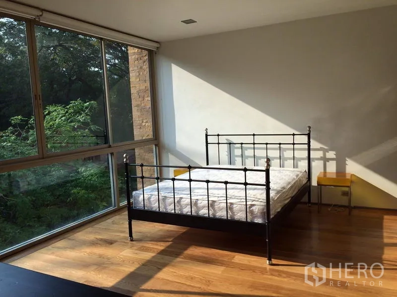Condominium for rent in Khlong Toei, Bangkok - Sunlit bedroom with metal bedframe, hardwood floors, and full-height corner windows facing greenery.