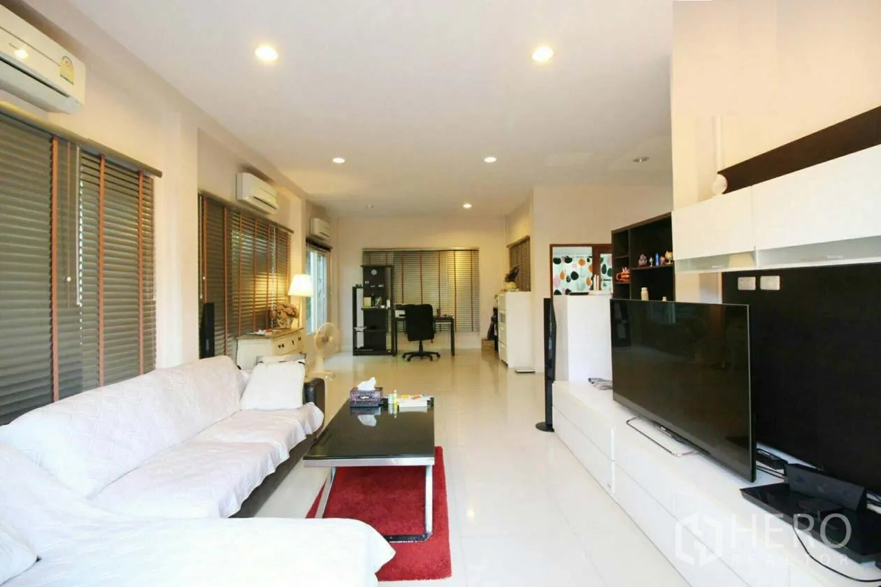 Detached House for rent or sale in Lat Krabang, Bangkok - Living area with sofa set, large TV, study nook and window blinds under recessed lighting.