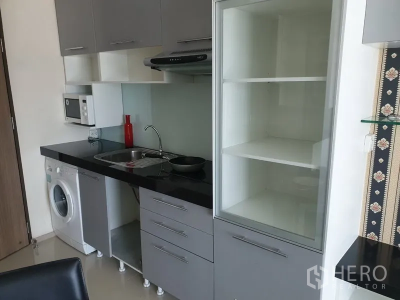 Condominium for rent in Khlong Toei, Bangkok - Compact modern kitchen with black countertops, sink, range hood, microwave, shelving, and in-unit washing machine.