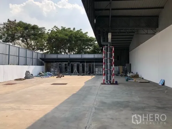 คลังสินค้า ให้เช่าในPak Kret, Nonthaburi - ลานคอนกรีตกว้างพร้อมหลังคาบางส่วน รั้วรอบขอบชิดและต้นไม้ด้านหลัง