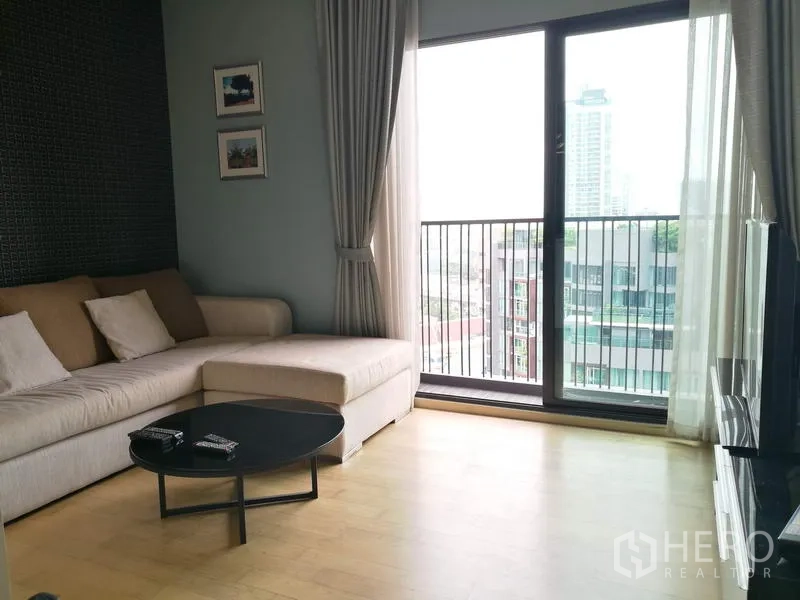 Condominium for rent in Watthana, Bangkok - Living room with L-shaped sofa and large sliding doors opening to a balcony with urban views.