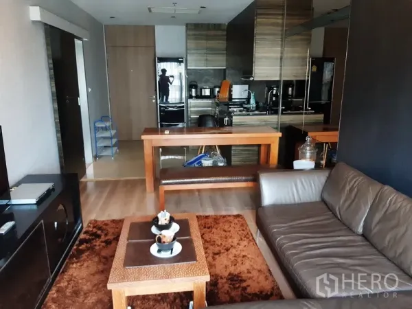 公寓 出租在Bangkok - 开放式起居区，面向带木质橱柜的现代小厨房，设用餐吧台与镜面墙。